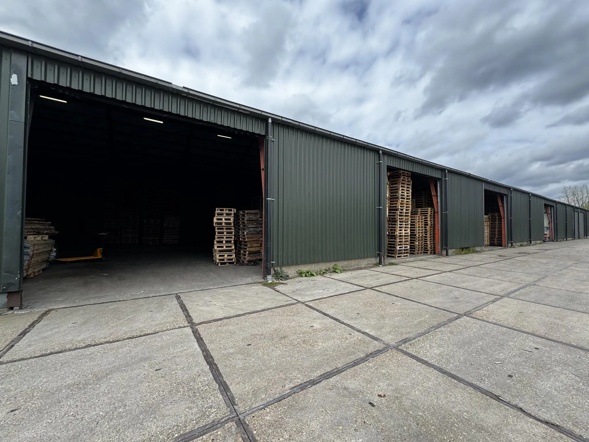 Rij donkere opslagloodsen aan de Graaf van Lynden van Sandenburgweg met stapels houten pallets bij de open ingangen.